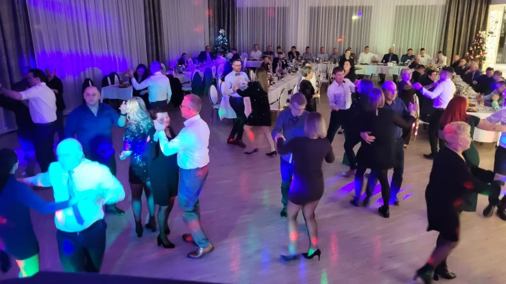 Duża grupa pracowników integruje się na parkiecie w przestronnej, nowoczesnej sali podczas imprezy firmowej z DJ-em.