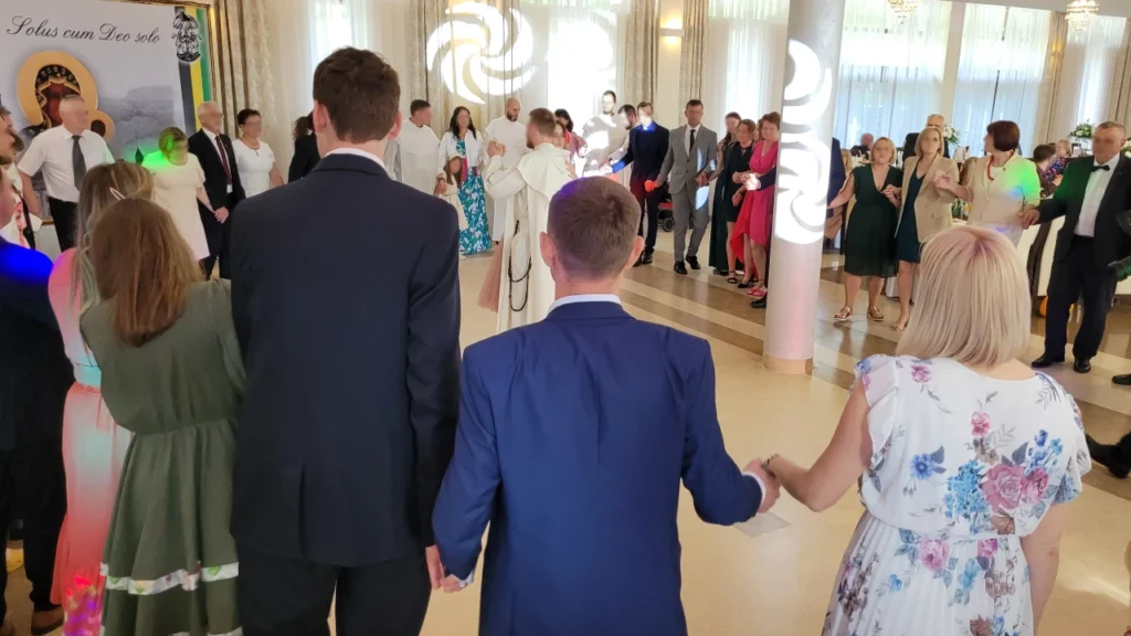 Ksiądz Prymicjant i goście trzymają się za ręce w dużym kole podczas zabawy integracyjnej na sali bankietowej.